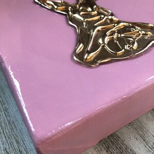 May include: A pink canvas with a gold-colored, three-dimensional sculpture of a woman in a flowing dress.