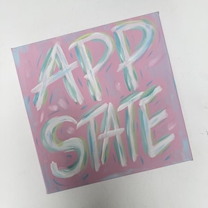 Op de afbeelding: Een vierkant canvas schilderij met een oudroze achtergrond en de tekst APP STATE geschilderd in grote, gelaagde penseelstreken van wit, lichtblauw en lichtgroen. De belettering heeft een getextureerd, handgeschilderd uiterlijk met zichtbare streken.