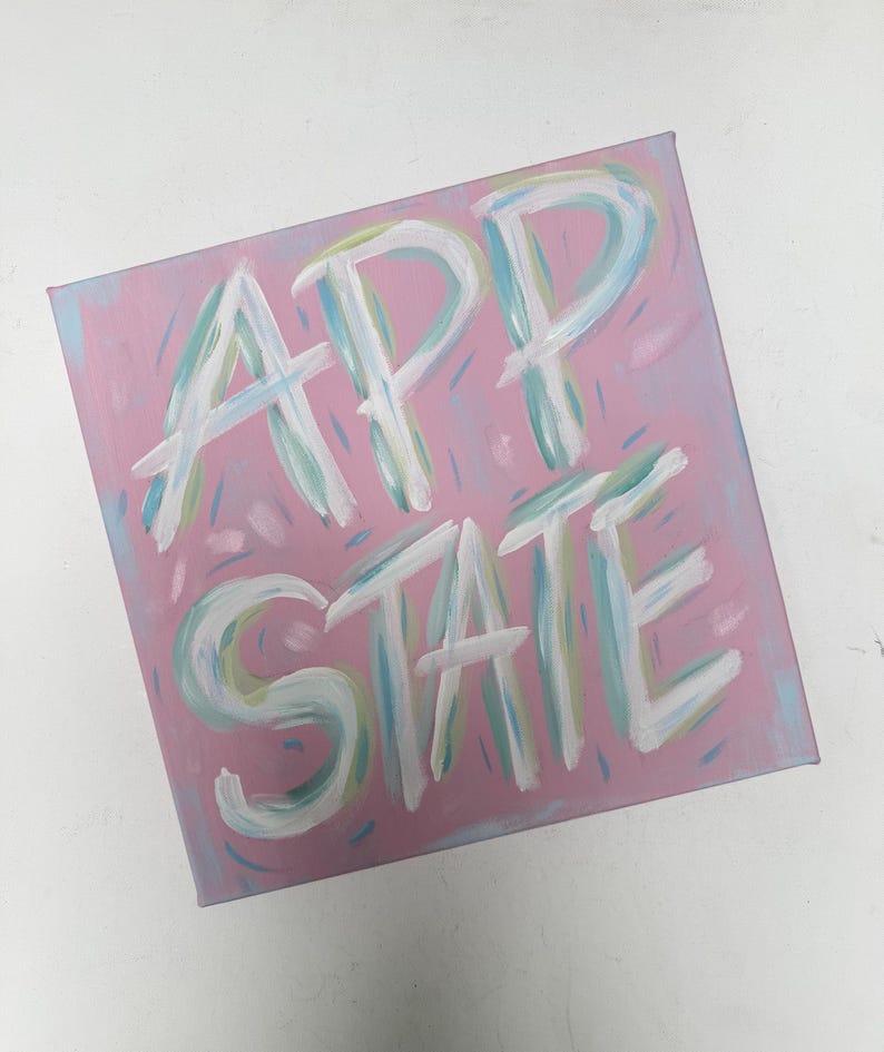 Op de afbeelding: Een vierkant canvas schilderij met een oudroze achtergrond en de tekst APP STATE geschilderd in grote, gelaagde penseelstreken van wit, lichtblauw en lichtgroen. De belettering heeft een getextureerd, handgeschilderd uiterlijk met zichtbare streken.