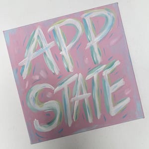 Peut inclure: Une peinture sur toile carr&eacute;e pr&eacute;sentant le texte APP STATE en grandes lettres blanches peintes &agrave; la main avec des accents de coups de pinceau bleu clair et vert. Le fond est rose clair uni, d&eacute;cor&eacute; de petits tirets dispers&eacute;s en bleu et vert.