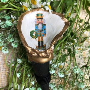 May include: A gold and white painted seashell bottle stopper with a nutcracker design. The nutcracker is wearing a blue and gold uniform and holding a green wreath.