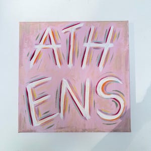 Op de afbeelding: Een vierkant canvas schilderij met het woord ATHENS in dikke witte letters. De achtergrond is zachtroze en de letters zijn omlijnd met levendige streken oranje, magenta en blauwgroene verf, wat een gelaagd en getextureerd effect geeft.