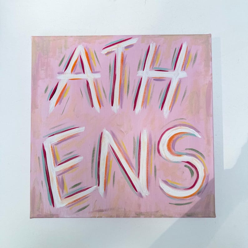 Op de afbeelding: Een vierkant canvas schilderij met het woord ATHENS in dikke witte letters. De achtergrond is zachtroze en de letters zijn omlijnd met levendige streken oranje, magenta en blauwgroene verf, wat een gelaagd en getextureerd effect geeft.