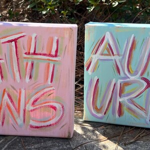 Peut inclure: Deux toiles carr&eacute;es avec des lettres peintes &agrave; la main. La toile rose affiche le mot ATHENS en coups de pinceau blancs, rouges et orange. La toile bleu clair affiche le mot AUBURN en coups de pinceau blancs, rouges et violets.