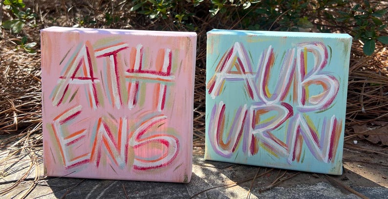 Peut inclure: Deux toiles carr&eacute;es avec des lettres peintes &agrave; la main. La toile rose affiche le mot ATHENS en coups de pinceau blancs, rouges et orange. La toile bleu clair affiche le mot AUBURN en coups de pinceau blancs, rouges et violets.