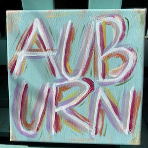 Peut inclure: Une peinture sur toile carr&eacute;e repr&eacute;sentant le mot AUBURN &eacute;crit en lettres blanches avec des coups de pinceau superpos&eacute;s de rose, violet, orange et jaune. Le fond est d'un bleu clair uni et les lettres ont un aspect textur&eacute; et peint &agrave; la main.