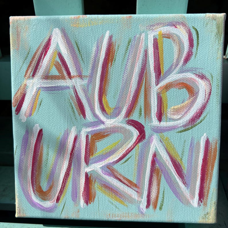 Peut inclure: Une peinture sur toile carr&eacute;e repr&eacute;sentant le mot AUBURN &eacute;crit en lettres blanches avec des coups de pinceau superpos&eacute;s de rose, violet, orange et jaune. Le fond est d'un bleu clair uni et les lettres ont un aspect textur&eacute; et peint &agrave; la main.