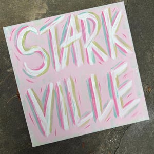 Peut inclure: Une peinture sur toile carr&eacute;e pr&eacute;sentant le texte STARK VILLE en lettres blanches &eacute;paisses peintes &agrave; la main. Le fond est d'un rose clair doux, accentu&eacute; par des coups de pinceau vert menthe, rose vif et dor&eacute; mat sur toute la surface.