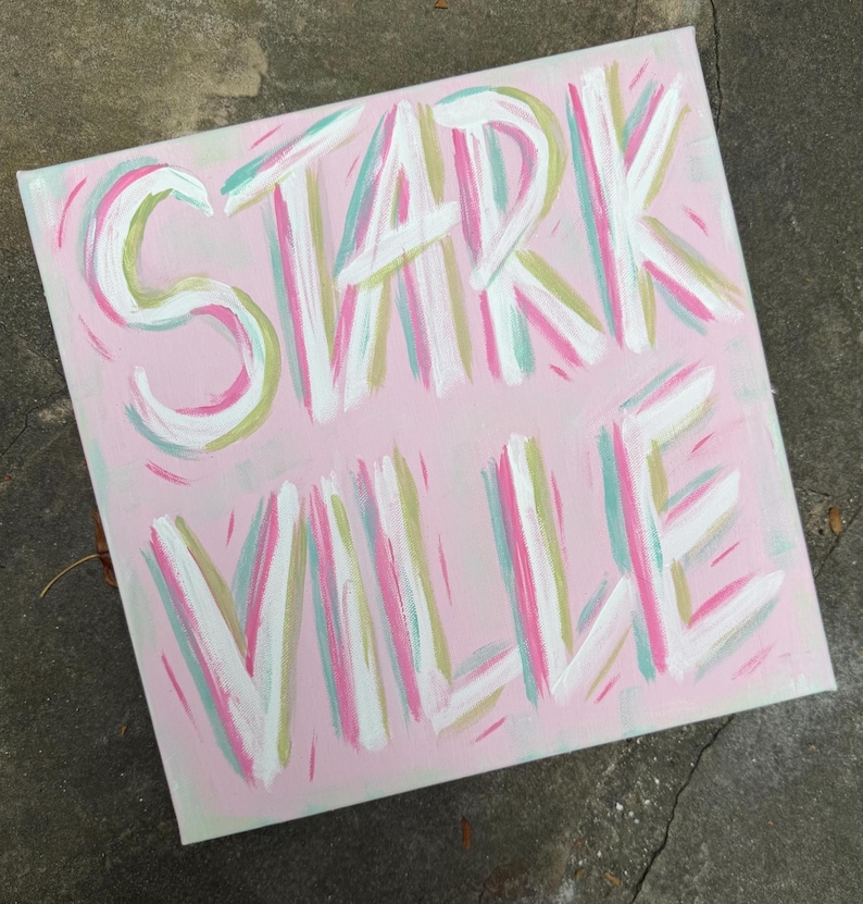 Peut inclure: Une peinture sur toile carr&eacute;e pr&eacute;sentant le texte STARK VILLE en lettres blanches &eacute;paisses peintes &agrave; la main. Le fond est d'un rose clair doux, accentu&eacute; par des coups de pinceau vert menthe, rose vif et dor&eacute; mat sur toute la surface.