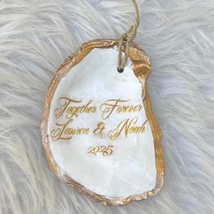 May include: A white oyster shell ornament with gold edging and a natural twine loop. The shell is inscribed with the words "Together Forever" and the names "Lauren & Noah" along with the year "2025" in gold script.