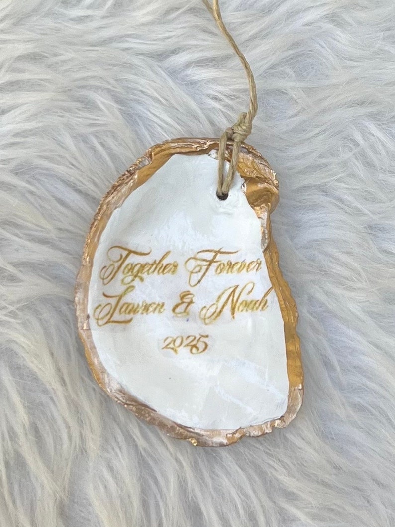 May include: A white oyster shell ornament with gold edging and a natural twine loop. The shell is inscribed with the words "Together Forever" and the names "Lauren & Noah" along with the year "2025" in gold script.