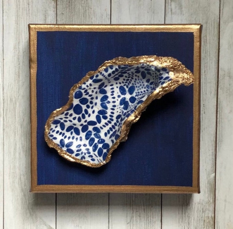 May include: A blue and white oyster shell with a gold rim, mounted on a blue canvas with a gold frame.