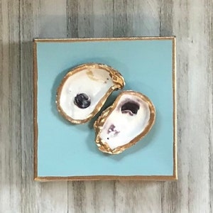 May include: Two oyster shells painted gold on a light blue background. The shells are arranged in a heart shape.