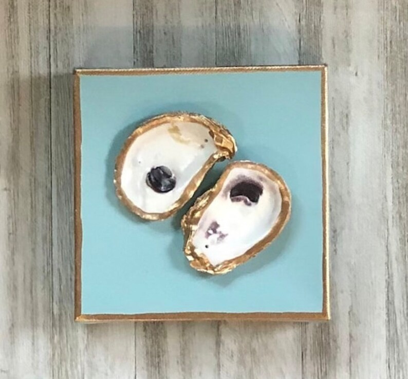 May include: Two oyster shells painted gold on a light blue background. The shells are arranged in a heart shape.