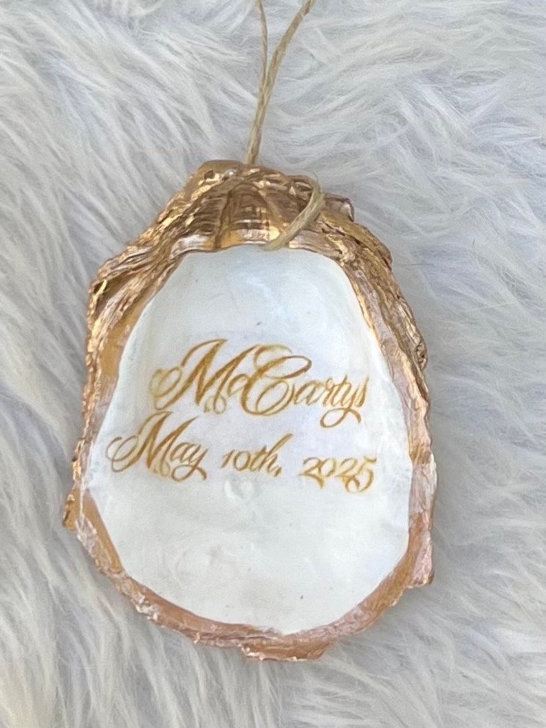May include: An oyster shell ornament with gold edging and the inscription "McCartys May 10th, 2025" in gold script. The shell's interior is white, with a natural texture. A twine loop is attached for hanging.