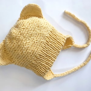 May include: A yellow knitted baby hat with bear ears and ties. The hat is made of a soft, textured yarn.