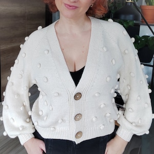 May include: A white knitted cardigan with a button closure and a textured bobble design. The cardigan has long sleeves and a relaxed fit.