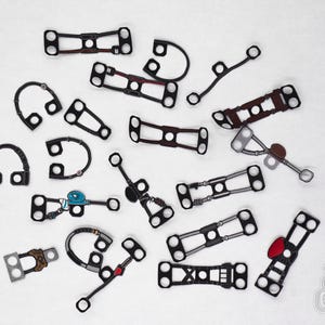 May include: A collection of 18 black and brown metal clasps with various shapes and designs. Some clasps have a red or blue accent. The clasps are arranged on a white surface.