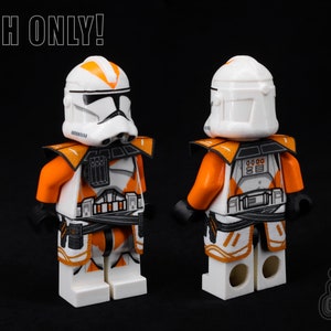Puede incluir: Una minifigura LEGO personalizada de un clon de soldado de Star Wars con un esquema de color naranja y blanco. La figura lleva un casco con un diseño blanco y naranja y tiene una pieza de pecho negra y naranja. La figura sostiene un rifle bláster en su mano derecha. "CLOTH ONLY!" está escrito en texto negro sobre un fondo blanco en la parte superior de la imagen.
