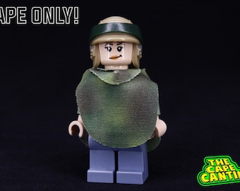 May include: A green and brown camouflage cape for a Lego minifigure. The cape is made of fabric and has a textured surface. The text "CAPE ONLY!" is visible in the top left corner of the image.
