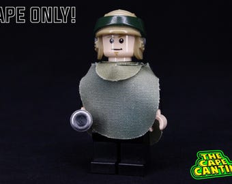 May include: A green and brown LEGO minifigure wearing a green cape and a tan helmet with a green visor. The minifigure is holding a black and silver weapon. The text "CAPE ONLY!" is visible in the top left corner of the image.