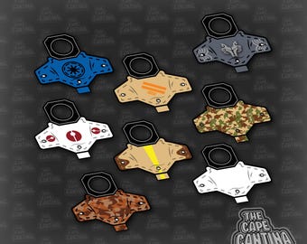 May include: A collection of eight flat, stylized cape accessories with circular neck openings, featuring various colors and patterns including blue, tan, camouflage, white, and brown, with the text THE CAPE CANTINA visible in the bottom right corner.