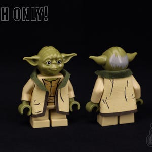 Puede incluir: Una minifigura Lego de Yoda verde y marrón que lleva una túnica marrón con un cuello verde. La minifigura está de pie sobre una superficie negra. "CLOTH ONLY!" está escrito en blanco en la parte superior de la imagen.
