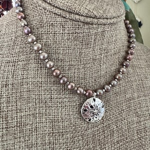 Puede incluir: Un collar con perlas marrón-grisáceas y un colgante de estrella de mar plateado. Las perlas varían en tamaño, y el colgante tiene un diseño calado detallado. El collar se muestra sobre una superficie texturizada de tono neutro.