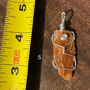 Peut inclure: Un pendentif en fil de fer avec un cristal naturel brun orang&eacute;. Le cristal mesure environ 5 cm de long et a une surface rugueuse et textur&eacute;e. Le fil est de couleur argent et est enroul&eacute; autour du cristal selon un motif g&eacute;om&eacute;trique.