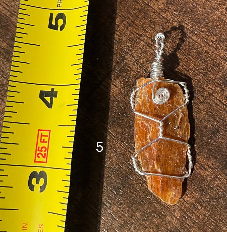 Peut inclure: Un pendentif en fil de fer avec un cristal naturel brun orang&eacute;. Le cristal mesure environ 5 cm de long et a une surface rugueuse et textur&eacute;e. Le fil est de couleur argent et est enroul&eacute; autour du cristal selon un motif g&eacute;om&eacute;trique.