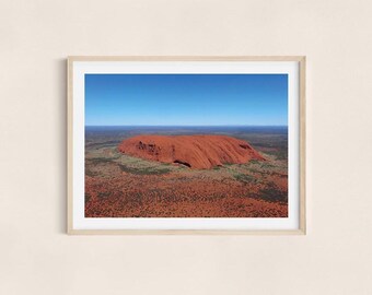 Op de afbeelding: Een ingelijste prent met een landschapsbeeld van Uluru, een grote zandsteenformatie in Australië. De afbeelding toont de rode rots onder een heldere blauwe lucht, omgeven door dor terrein.
