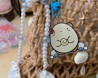 May include: A beige macrame bag with a blue clasp, pearl beaded accents, and seashell charms. A cartoon character keychain with a flower and seashell details hangs from the bag. The bag is on a light brown wooden surface.
