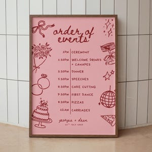 May include: A framed pink wedding schedule with a wooden border. The sign displays the words "order of events" and a detailed timeline of the day's activities, accompanied by illustrations. The date "11th July 2025" is also present.