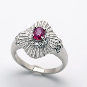 Könnte beinhalten: Ein silberner Ring mit einem großen, ovalen Rubin, der von kleineren Diamanten umgeben ist. Die Ringschiene ist texturiert und hat ein fächerförmiges Design.
