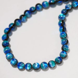 Puede incluir: Un collar hecho de cuentas de vidrio azul y negro. Las cuentas tienen un acabado moteado e iridiscente.