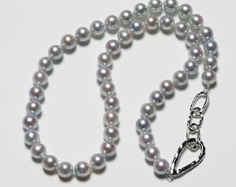 May include: A strand of lustrous, round, gray pearls. The pearls are strung together, forming a necklace. The necklace is fastened with a silver-colored clasp. The pearls have a subtle iridescence, reflecting light. The necklace is displayed on a white surface.