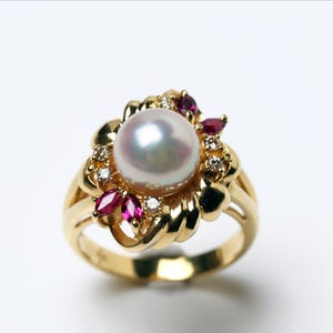May include: A gold ring featuring a large, lustrous pearl at the center, surrounded by smaller rubies and diamonds. The ring's setting has a floral design. The band is a polished gold. A beautiful piece of jewelry.