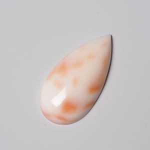 Op de afbeelding: Een traanvormige cabochon van witte en oranje schelp met een glad, gepolijst oppervlak.