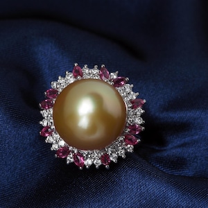 May include: A gold-toned pearl ring with a halo of pink gemstones and white diamonds. The ring's central pearl is surrounded by a cluster of smaller, vibrant pink stones and sparkling white diamonds, set against a dark blue background.