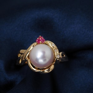 May include: A gold ring featuring a large, lustrous white pearl as the centerpiece. The ring is adorned with small red gemstones and clear accent stones. The band has an intricate, swirling design.
