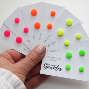 May include: A set of six pairs of small, round stud earrings in bright neon colours: pink, orange, and green. The earrings are displayed on white cards with the text "You were born to wear Sparkles".