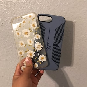 May include: Two phone cases, one clear with a white daisy pattern and the other a solid blue with a textured design and the word "neill" printed on it.