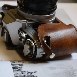 Op de afbeelding: Een zilveren Canon camera met een bruine leren cameratas. De camera heeft een zwarte lens en een zilveren wijzerplaat met cijfers.