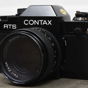 Può includere: Una fotocamera Contax RTS nera con un obiettivo Carl Zeiss Planar. La fotocamera ha una tracolla in pelle nera.