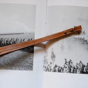 Puede incluir: Una correa de cuero marrón con un anillo de metal plateado y dos remaches de latón. La correa está sobre un libro con una fotografía en blanco y negro de un grupo de personas.