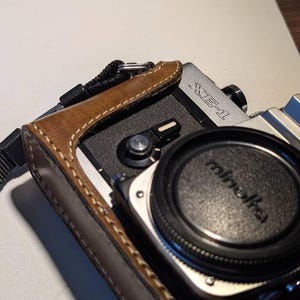 Könnte beinhalten: Eine Vintage Minolta XE-1 Kamera mit schwarzem Objektivdeckel und brauner Ledertasche. Das Kameragehäuse ist silberfarben mit schwarzen Akzenten. Die Ledertasche hat weiße Nähte und einen schwarzen Riemen.