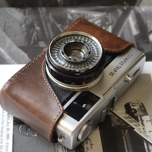 Op de afbeelding: Een zilveren Olympus Trip 35 camera met een zwarte lens en een bruine leren hoes. De camera ligt op een bedrukte pagina met zwart-wit afbeeldingen en tekst. De lens is scherp en de camerabody is schoon.