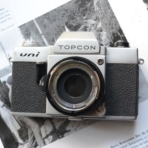 Può includere: Una macchina fotografica a pellicola Topcon Uni 35 mm di colore argento e nero con un tappo dell'obiettivo sull'obiettivo. La macchina fotografica ha un corpo nero testurizzato e una piastra superiore argentata con le parole "TOPCON" e "uni" stampate su di essa.