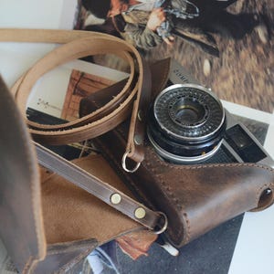 May include: A vintage camera with a leather case and strap. The camera is silver and black, with a prominent lens. The brown leather case is open, revealing the camera. The strap is also brown, and the scene is set on a surface with printed images.