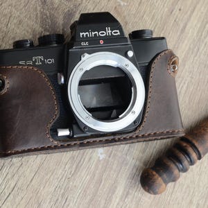 Può includere: Un corpo macchina fotografica a pellicola Minolta SRT 101 nero con innesto obiettivo argentato, recante la scritta Minolta e SRT 101, inserito in una mezza custodia in pelle marrone scuro con cuciture visibili, accanto a un brunitoio in legno.